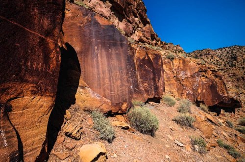 Petroglyphs etched into a rocky cliffside, surrounded by arid terrain and sparse vegetation under a clear blue sky.