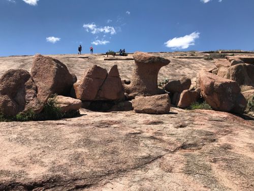 Rocky landscape with large boulders and a flat surface, under a blue sky with a few clouds and distant figures on top.