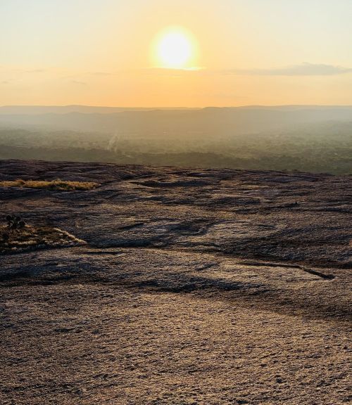 Sunset over a rocky landscape, with soft hills and a hazy horizon in the background.