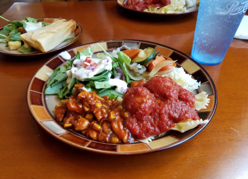 A colorful plate with pasta, chili, salad, rice, and a drink, set on a wooden table.