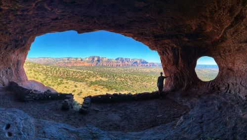 A person stands inside a cave, looking out at a panoramic view of mountains and desert landscape through two openings.