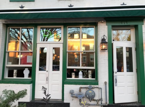 Exterior of a building with large windows, green trim, and two white doors, featuring a gas meter and plants nearby.