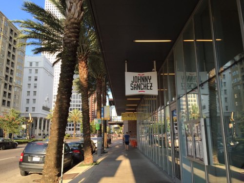 A city sidewalk lined with palm trees, featuring a restaurant sign for "Johnny Sanchez" and nearby buildings.