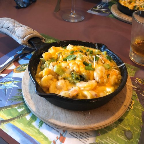 A cast iron skillet filled with cheesy macaroni, shrimp, broccoli, and green peppers, served on a wooden board.