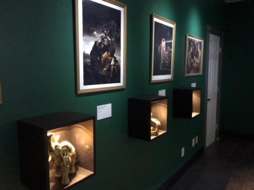 A dimly lit gallery with green walls, featuring framed paintings and illuminated display cases with elephant sculptures.
