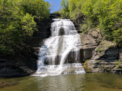 A cascading waterfall surrounded by lush green trees and rocky cliffs, reflecting sunlight in a serene natural setting.