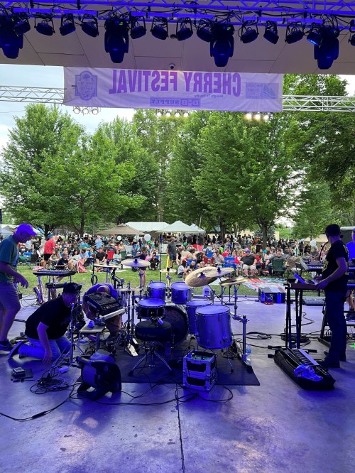 A stage setup with drums and instruments, overlooking a crowd at a festival under trees.