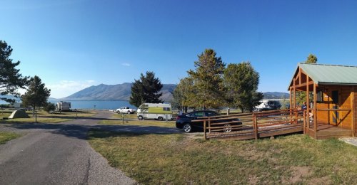 Scenic lakeside view with trees, parked vehicles, and a cabin against a backdrop of mountains and clear blue sky.