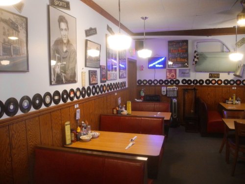 A cozy diner with wooden walls, vinyl records lining the walls, and booths set for dining. Elvis Presley photo on display.