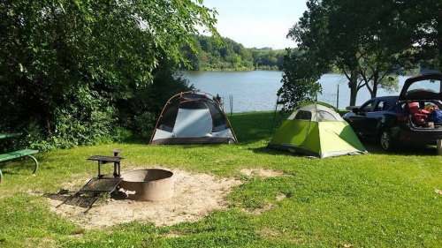Two tents set up by a lake, with a fire pit and a car nearby, surrounded by greenery and a clear blue sky.