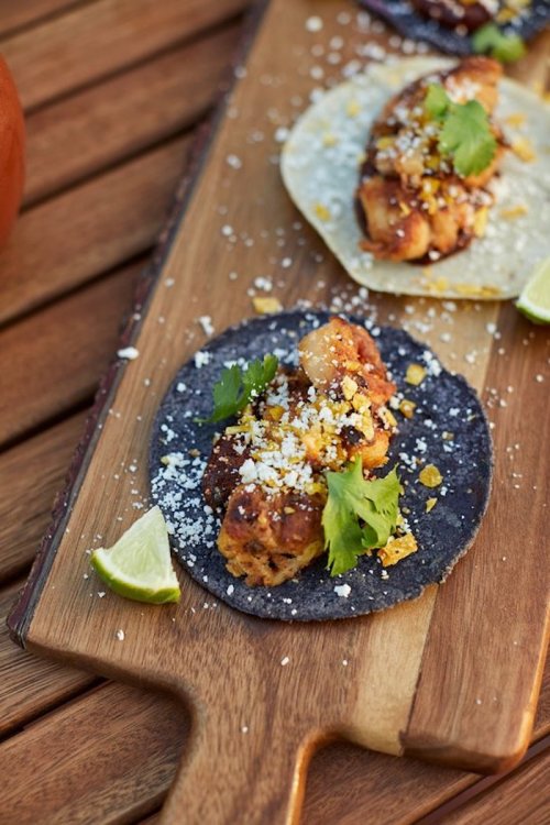 A wooden board with blue corn tacos topped with grilled chicken, cheese, corn, cilantro, and lime wedges.