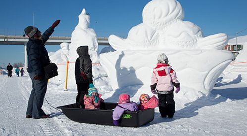 A group of children in winter clothing admire large snow sculptures while one adult gestures towards them.