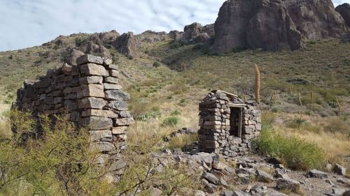 Ruins of stone structures in a desert landscape, surrounded by rocky hills and sparse vegetation under a cloudy sky.