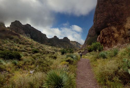 A hiker walks along a dirt path through a lush valley surrounded by rocky mountains and cloudy skies.