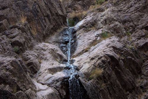 A small waterfall cascading down rocky terrain, surrounded by sparse vegetation and rugged cliffs.