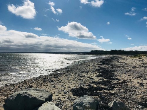 A rocky beach under a bright sky with scattered clouds, reflecting sunlight on the water's surface.