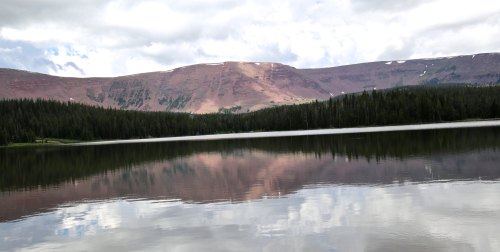 A serene lake reflects mountains and clouds, surrounded by lush green trees under a partly cloudy sky.