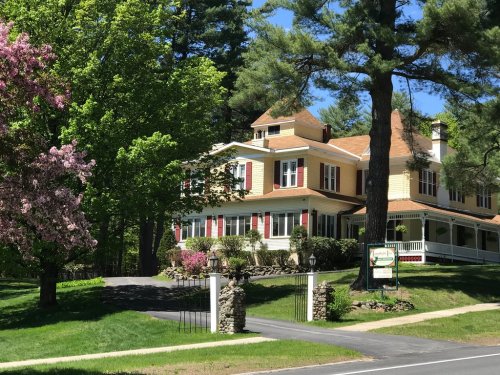 A large yellow house with a porch, surrounded by green trees and blooming flowers on a sunny day.