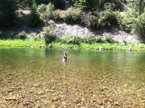A person stands in a clear river, arms raised, surrounded by lush greenery and rocky banks.