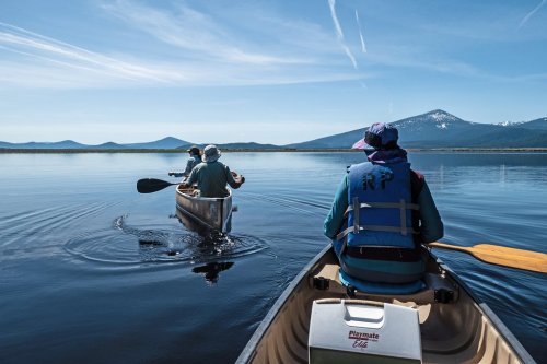 Two people canoeing on a calm lake with mountains in the background under a clear blue sky.
