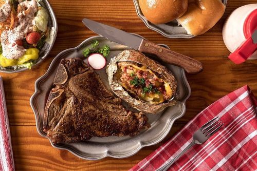 A plate with a large steak, a stuffed baked potato, and a side salad, accompanied by rolls and a drink.