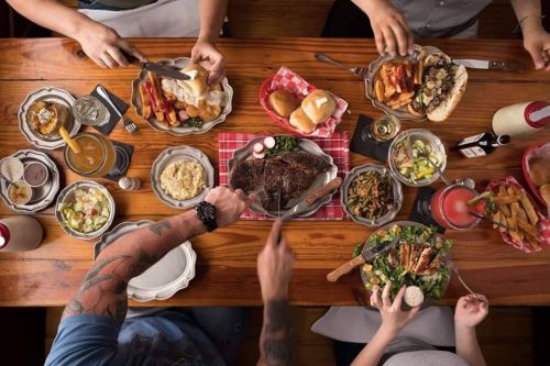 A table filled with various dishes, including steak, salads, and sides, as people enjoy a meal together.
