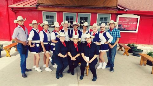 A group of dancers in cowboy hats and outfits poses together outside a red building, smiling for the camera.