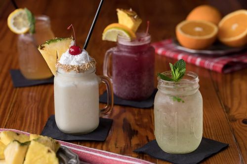 Four colorful cocktails in mason jars, garnished with fruit and mint, on a wooden table with fresh pineapple slices.