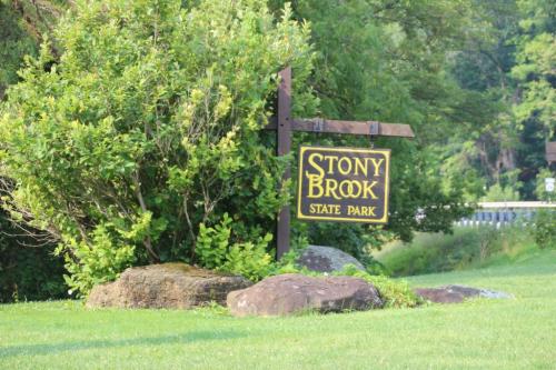 Sign for Stony Brook State Park surrounded by greenery and rocks, with a road visible in the background.