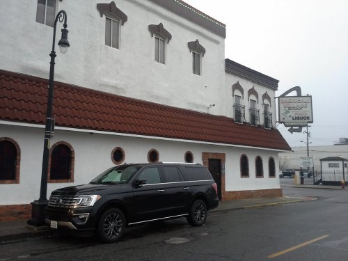 A black SUV parked in front of a white building with a red-tiled roof and a liquor store sign on a rainy day.