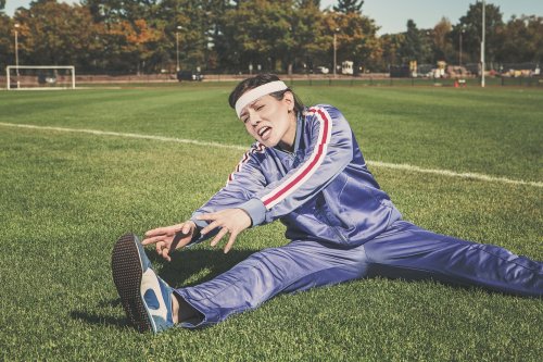 A person in a blue tracksuit stretches on a grassy field, wearing a headband and showing a focused expression.