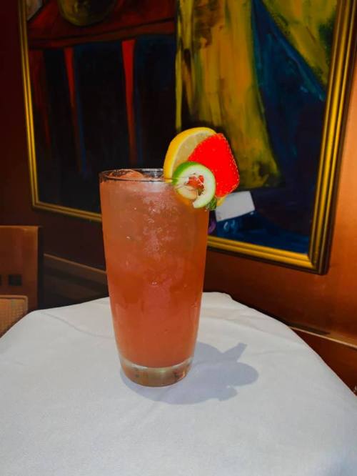 A tall glass of colorful cocktail garnished with lemon, strawberry, and kiwi, set on a white tablecloth.