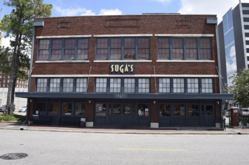 A brick building with large windows and a sign reading "Suga's," located on a city street.