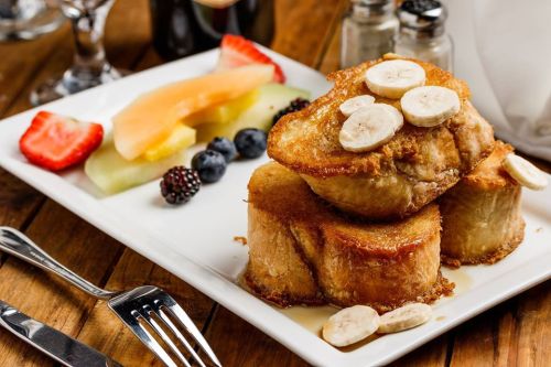 A plate of French toast topped with banana slices, served with a side of fresh fruit including berries and melon.