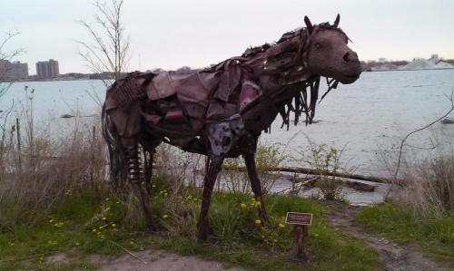 A sculpture of a horse made from metal and scrap materials, standing near a body of water with greenery around it.