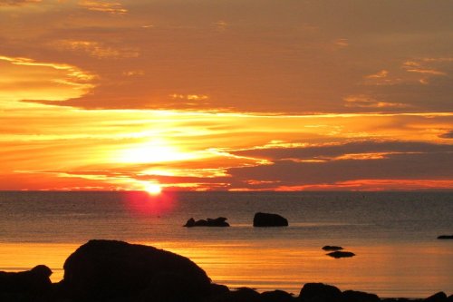 A serene sunset over calm waters, with silhouettes of rocks in the foreground and vibrant orange and yellow hues in the sky.