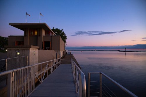 A modern building by the water at dusk, with a wooden walkway leading to it and a calm, reflective surface.