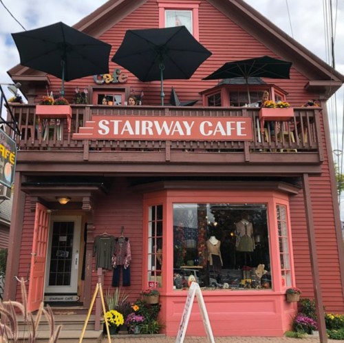 A charming red café with a sign reading "Stairway Café," featuring outdoor umbrellas and colorful flower pots.