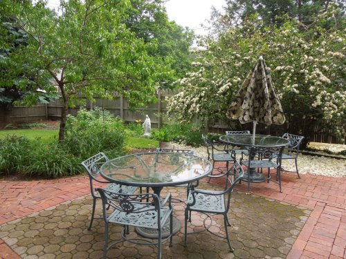 A serene backyard patio with a glass table, metal chairs, and a large umbrella surrounded by greenery and flowers.