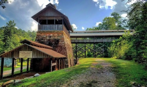 Historic building with a wooden structure above, surrounded by trees and a gravel path under a blue sky with clouds.