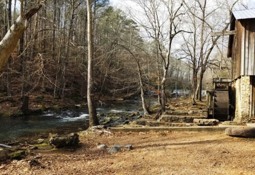 A serene riverside scene featuring a wooden structure and a waterwheel, surrounded by bare trees and rocky terrain.