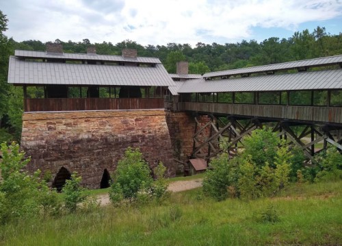 A modern wooden structure with a sloped roof, built over a stone foundation, surrounded by greenery and trees.