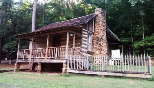 A rustic wooden cabin with a stone chimney, surrounded by trees and a grassy area.