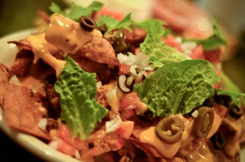 A close-up of a plate of nachos topped with cheese, olives, tomatoes, onions, and fresh green leaves.