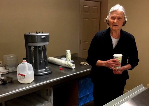 An elderly woman stands at a kitchen counter holding a cup, with a milk jug and a vintage mixer nearby.