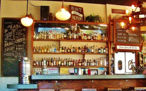 A rustic bar with shelves filled with various liquor bottles, a chalkboard menu, and a cozy atmosphere.