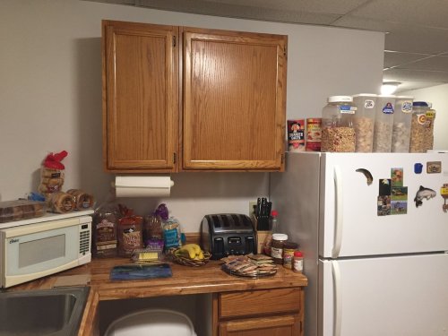 A kitchen corner with a wooden countertop, microwave, toaster, snacks, and a fridge topped with jars of cereal.