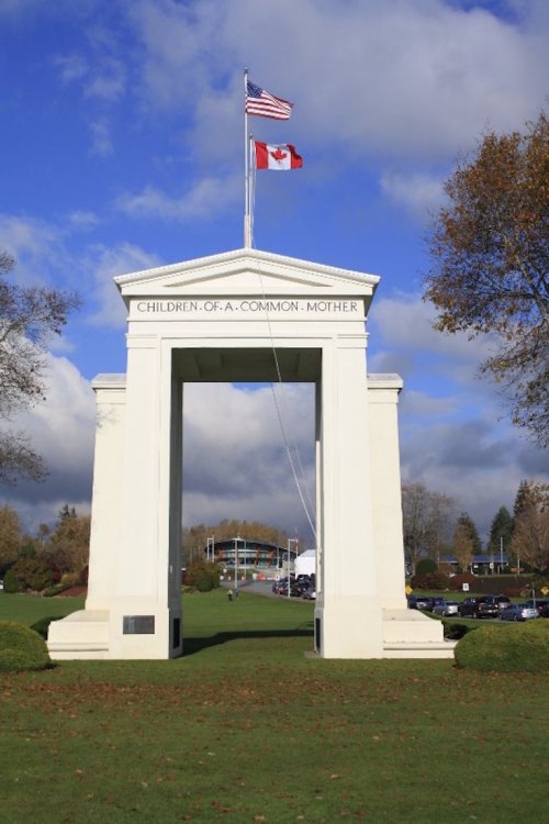 White archway with flags of the U.S. and Canada, surrounded by green grass and trees under a blue sky.