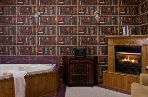 Cozy bathroom with a vintage typewriter, fireplace, and bookshelf wallpaper, featuring a jacuzzi tub and warm decor.