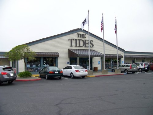 Exterior of a building labeled "The Tides," with flags and parked cars in a lot on a cloudy day.